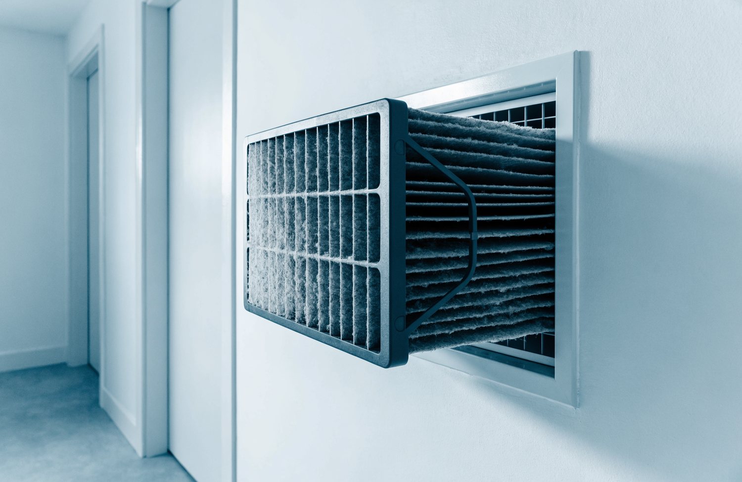 A pleated furnace filter pulled halfway out of a wall return vent, pleats loaded with grey dust.